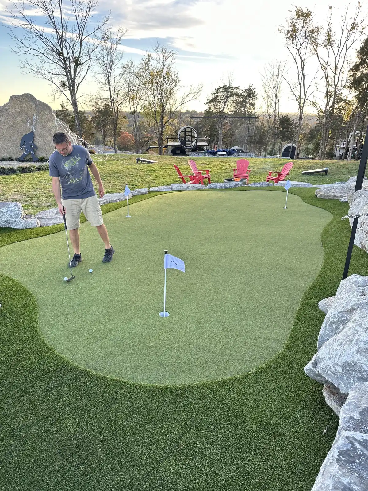 Man practicing putting on a custom contoured green with flags, boulder edging, and Adirondack chairs