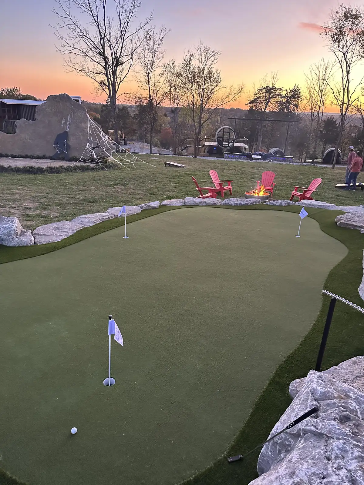 Custom backyard putting green at sunset with flag pins and boulder borders